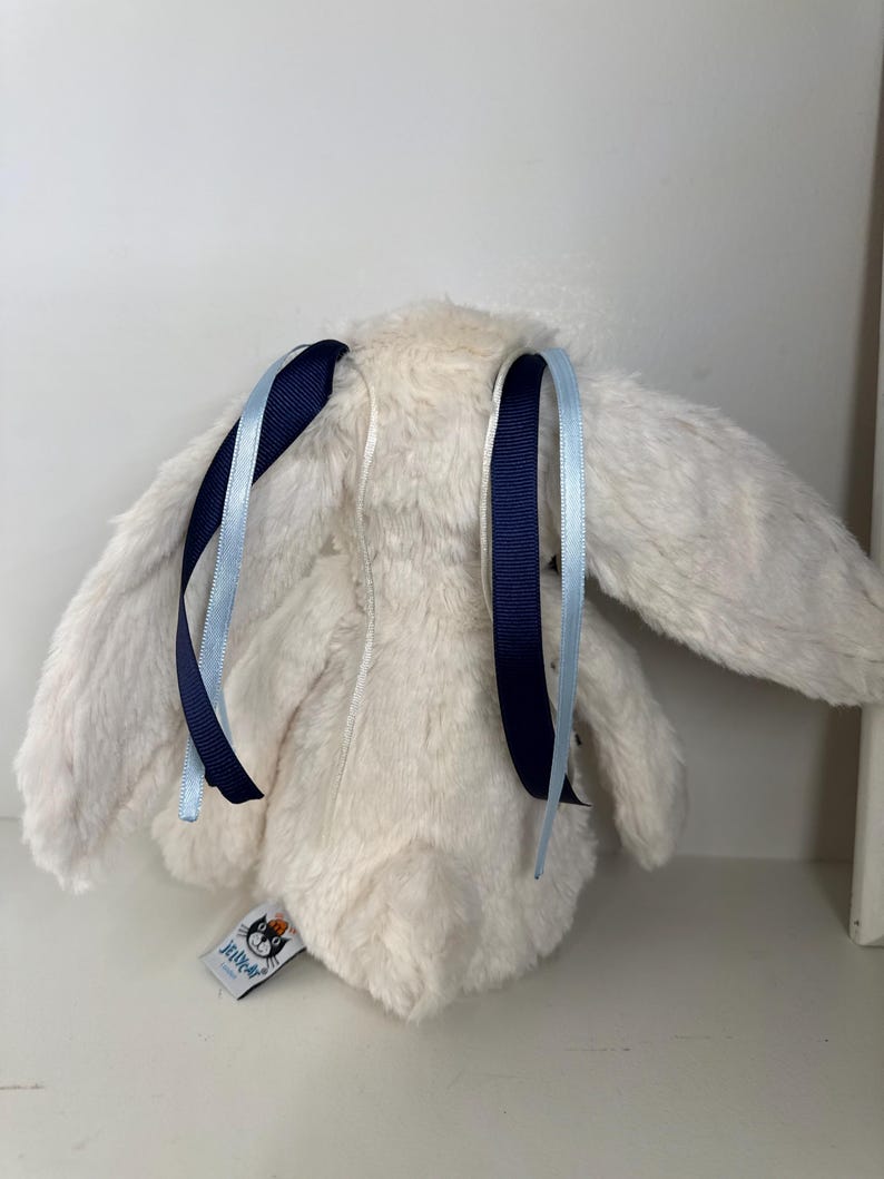 May include: A plush, cream-colored bunny toy with long ears and a fluffy texture. The bunny has dark blue and light blue ribbons tied to its ears. A small tag with a logo is attached to the bunny's body.