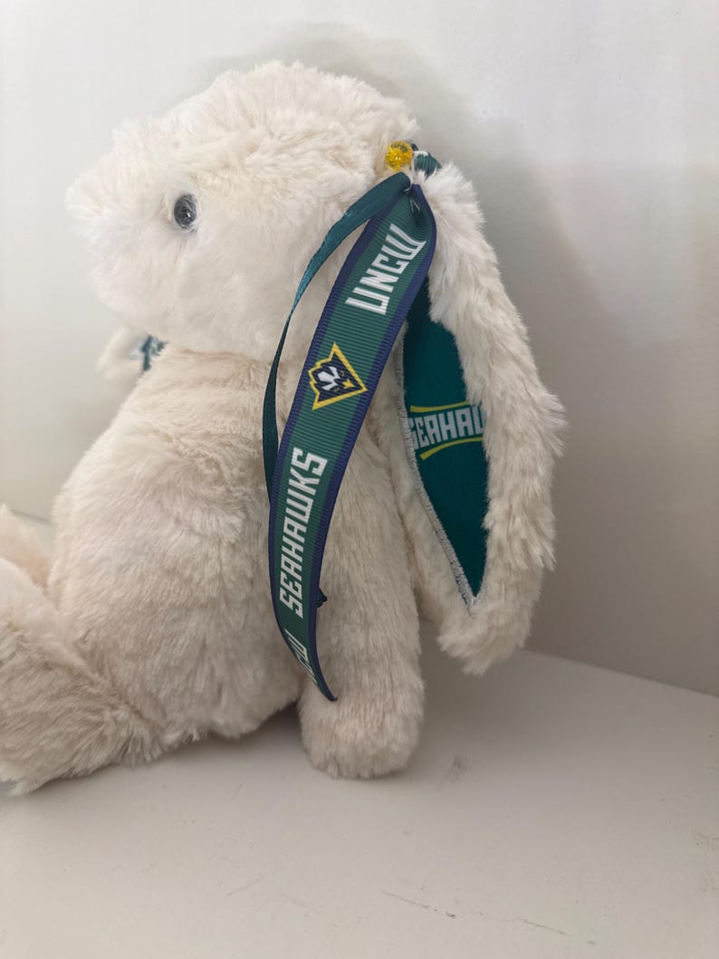 May include: A plush, cream-colored bunny with long ears. The bunny has a tag with the text "UNCW SEAHAWKS" in white lettering on a dark green ribbon. The bunny's eyes are black, and it has a soft, fluffy texture.