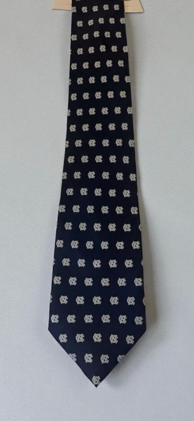 May include: Navy blue tie with a repeating pattern of white interlocking "NC" logos. The tie is displayed against a white background, showcasing its design. A classic accessory for formal or semi-formal attire.