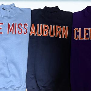May include: Five sweatshirts in various colors, each with a different college name in bold letters. The colors include blue with "DUKE", light blue with "OLE MISS", navy with "AUBURN", purple with "CLEMSON", and red with "ALABAMA".