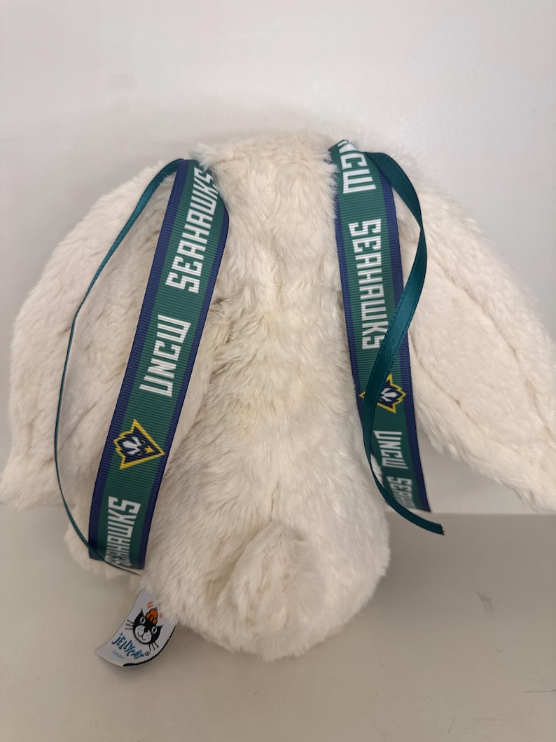 May include: A cream-colored plush bunny with long ears. The bunny has two green and navy blue ribbons attached to its back, each printed with "UNCW SEAHAWKS" and a logo. A small tag with a cat logo is attached to the bunny.