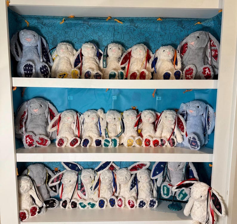 May include: A collection of plush bunny rabbits in a white display case. The bunnies are primarily white with various colored accents on their ears and feet. Some bunnies are gray or blue. The display case has three shelves and a blue patterned backdrop.