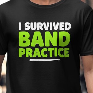 Può includere: T-shirt nero con la scritta "I Survived Band Practice" in bianco e verde. La maglietta ha un design semplice con una grafica di bacchetta. È una maglietta girocollo a maniche corte.