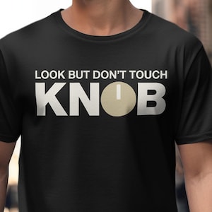 May include: Black t-shirt with the text "LOOK BUT DON'T TOUCH" above the word "KNOB". The word "KNOB" is in large, bold, white letters with a beige circle replacing the "O".