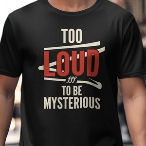 May include: A black short-sleeved t-shirt featuring the text "TOO LOUD TO BE MYSTERIOUS" printed on the front. The word "LOUD" is in large red capital letters, while the remaining text is in white capital letters with a white graphic line.