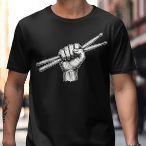 Può includere: T-shirt nero con grafica bianca di un pugno chiuso che tiene due bacchette. L'illustrazione dettagliata crea un forte impatto visivo. Questo design è ideale per batteristi e amanti della musica, offrendo una dichiarazione di stile audace.