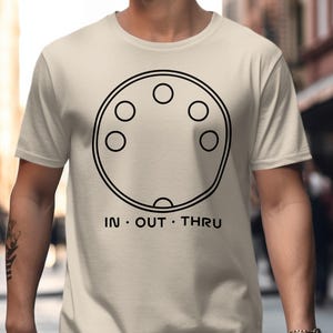 May include: Beige t-shirt with a black circular design featuring six circles and the words "IN • OUT • THRU" in black. The shirt is made of a soft material and is suitable for casual wear.