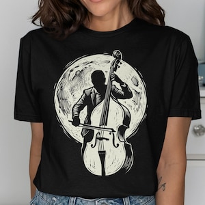 May include: A black short-sleeve t-shirt featuring a graphic print of a musician playing a double bass in front of a large, textured full moon. The illustration is rendered in cream and black ink, showcasing the silhouette of the player.