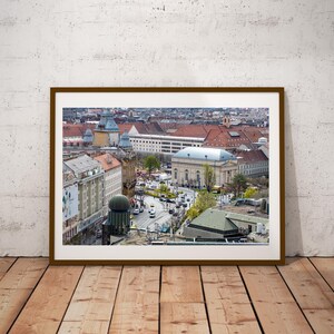 May include: A framed photograph of a cityscape, featuring buildings with red tile roofs, a domed structure, and a wide street with traffic. The image is set against a white wall and wooden floor.