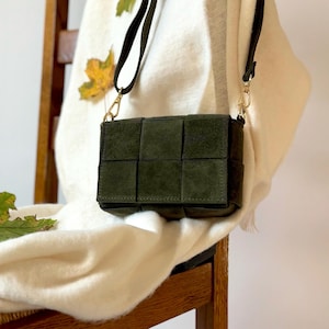 May include: A dark green suede crossbody bag with a woven design. The bag has a long, thin strap and gold-colored hardware. The bag is hanging on a wooden chair, with a white sweater in the background.