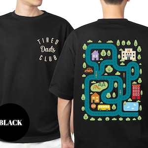 May include: Black t-shirt with "Tired Dads Club" text on the front and a colorful road map design on the back. The map includes houses, a hospital, a school, a bus, and a lake. The shirt is a casual wear item.