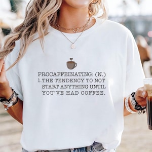 May include: White t-shirt with the definition of "Procaffeinating" printed in black. The definition includes a coffee cup graphic with a heart above it. The text reads: "The tendency to not start anything until you've had coffee."