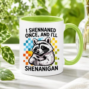 May include: A white ceramic mug with a light green interior and handle. The front features a graphic of a raccoon with black and white fur, surrounded by colorful pixelated squares. The text reads, I SHENNANED ONCE, AND I'LL SHENANIGAN.