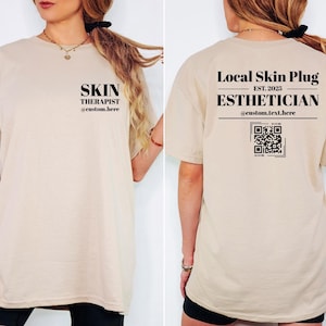 May include: A beige t-shirt with black text on the front that reads "SKIN THERAPIST @custom.here." The back of the shirt has the text "Local Skin Plug EST. 2023 ESTHETICIAN @custom.text.here" and a QR code. The shirt is short-sleeved.