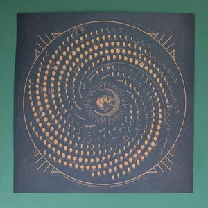 May include: A dark blue square wall calendar for the year 2027 featuring a spiral design of gold moon phases. The centre displays a detailed moon illustration surrounded by the months of the year, with small moon icons tracking daily lunar cycles.