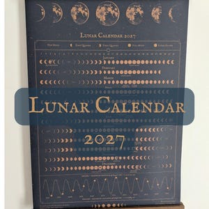 May include: A 2027 lunar calendar wall hanging featuring a dark blue background with copper-colored moon phases, text, and astronomical data. The print is held by two horizontal wooden bars at the top and bottom with a hanging cord.