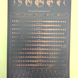 May include: A dark grey wall calendar titled Lunar Calendar 2027 with copper-coloured moon phase illustrations and text. The calendar is held by wooden magnetic rails at the top and bottom, featuring a monthly grid of moon cycles and a wave graph.