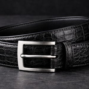 May include: A black leather belt with a textured, crocodile-like pattern. The belt features a silver-colored, rectangular buckle. The belt is coiled on a dark surface, showcasing its design and craftsmanship.