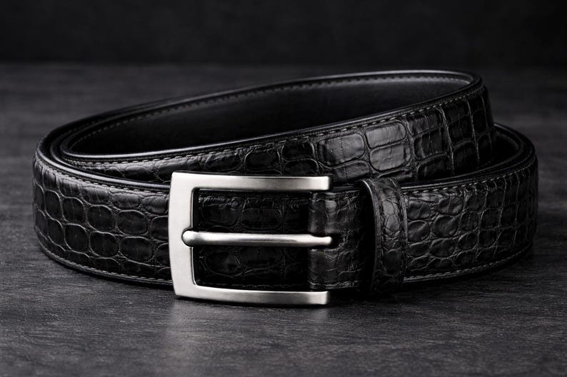 May include: A black leather belt with a textured, crocodile-like pattern. The belt features a silver-colored, rectangular buckle. The belt is coiled on a dark surface, showcasing its design and craftsmanship.