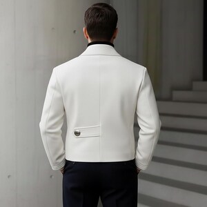 May include: A white jacket with a black turtleneck collar. The jacket has a single-button detail on the back and is paired with dark blue pants. The person is viewed from the back, showcasing the jacket's tailored design.