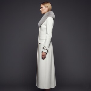May include: A long, white coat with a gray fur collar. The coat features a tailored design with a fitted waist, a small pocket, and a buckle detail on the sleeve. The coat is a classic outerwear piece.