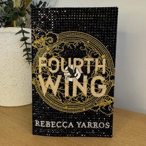 Peut inclure: Une couverture de livre noire avec le titre "FOURTH WING" en lettres dorées, entouré d'un motif doré. Le nom de l'auteur, "REBECCA YARROS", est en bas. La couverture est ornée de petits accents noirs et dorés scintillants.