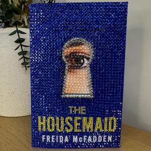 Puede incluir: Una cubierta de libro azul cubierta de brillantes pedrería. Presenta un diseño de ojo de cerradura con un ojo en su interior. El título "THE HOUSEMAID" y el nombre de la autora, Freida McFadden, están en letras doradas.