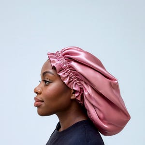 Puede incluir: Un gorro de satén rosa empolvado con un borde con volantes. El gorro está diseñado para proteger el cabello mientras se duerme. El tejido liso y brillante refleja la luz, creando reflejos y sombras. El gorro lo lleva una persona de piel oscura.