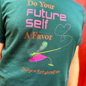 Puede incluir: Camiseta verde oscuro con el texto "Do Your Future Self A Favor" en naranja y rosa. Tambi&eacute;n presenta una ilustraci&oacute;n de lib&eacute;lula con un ala rosa y un dise&ntilde;o de coraz&oacute;n punteado. La camiseta tambi&eacute;n tiene el texto "You'll get to thank yourself later."