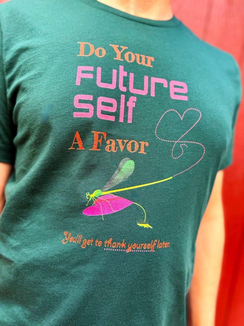 Puede incluir: Camiseta verde oscuro con el texto "Do Your Future Self A Favor" en naranja y rosa. Tambi&eacute;n presenta una ilustraci&oacute;n de lib&eacute;lula con un ala rosa y un dise&ntilde;o de coraz&oacute;n punteado. La camiseta tambi&eacute;n tiene el texto "You'll get to thank yourself later."