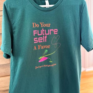 Puede incluir: Camiseta verde azulado con el texto rosa "Do Your Future Self A Favor" y una ilustraci&oacute;n de lib&eacute;lula. La camiseta tambi&eacute;n incluye la frase "You'll get to thank yourself later" en una fuente m&aacute;s peque&ntilde;a.