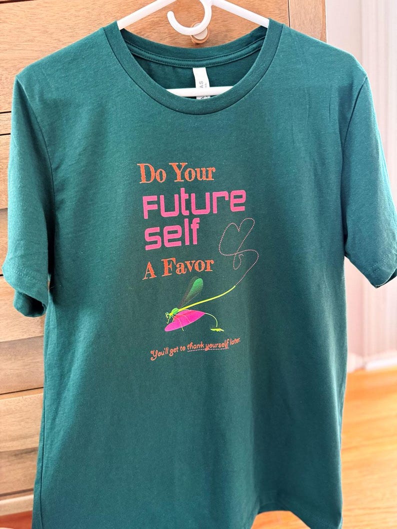 Puede incluir: Camiseta verde azulado con el texto rosa "Do Your Future Self A Favor" y una ilustraci&oacute;n de lib&eacute;lula. La camiseta tambi&eacute;n incluye la frase "You'll get to thank yourself later" en una fuente m&aacute;s peque&ntilde;a.