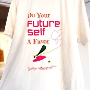 Puede incluir: Camiseta de color crema con el texto "Do Your Future Self A Favor" en rojo y naranja. Una ilustraci&oacute;n caprichosa de una lib&eacute;lula con detalles rosas y verdes se encuentra debajo del texto. La frase "You'll get to thank yourself later" tambi&eacute;n est&aacute; impresa.