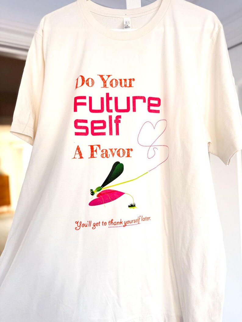 Puede incluir: Camiseta de color crema con el texto "Do Your Future Self A Favor" en rojo y naranja. Una ilustraci&oacute;n caprichosa de una lib&eacute;lula con detalles rosas y verdes se encuentra debajo del texto. La frase "You'll get to thank yourself later" tambi&eacute;n est&aacute; impresa.