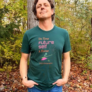 Puede incluir: Una camiseta verde oscuro con el texto "Do Your Future Self A Favor" en rosa y blanco. La camiseta presenta un gr&aacute;fico de una lib&eacute;lula y las palabras "You'll get to thank yourself later."