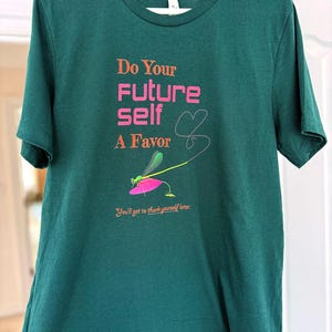 Puede incluir: Una camiseta verde oscuro con el texto "Do Your Future Self A Favor" en naranja y rosa. Un gr&aacute;fico de lib&eacute;lula est&aacute; debajo del texto, y la frase "You'll get to thank yourself later" est&aacute; impresa en la parte inferior.