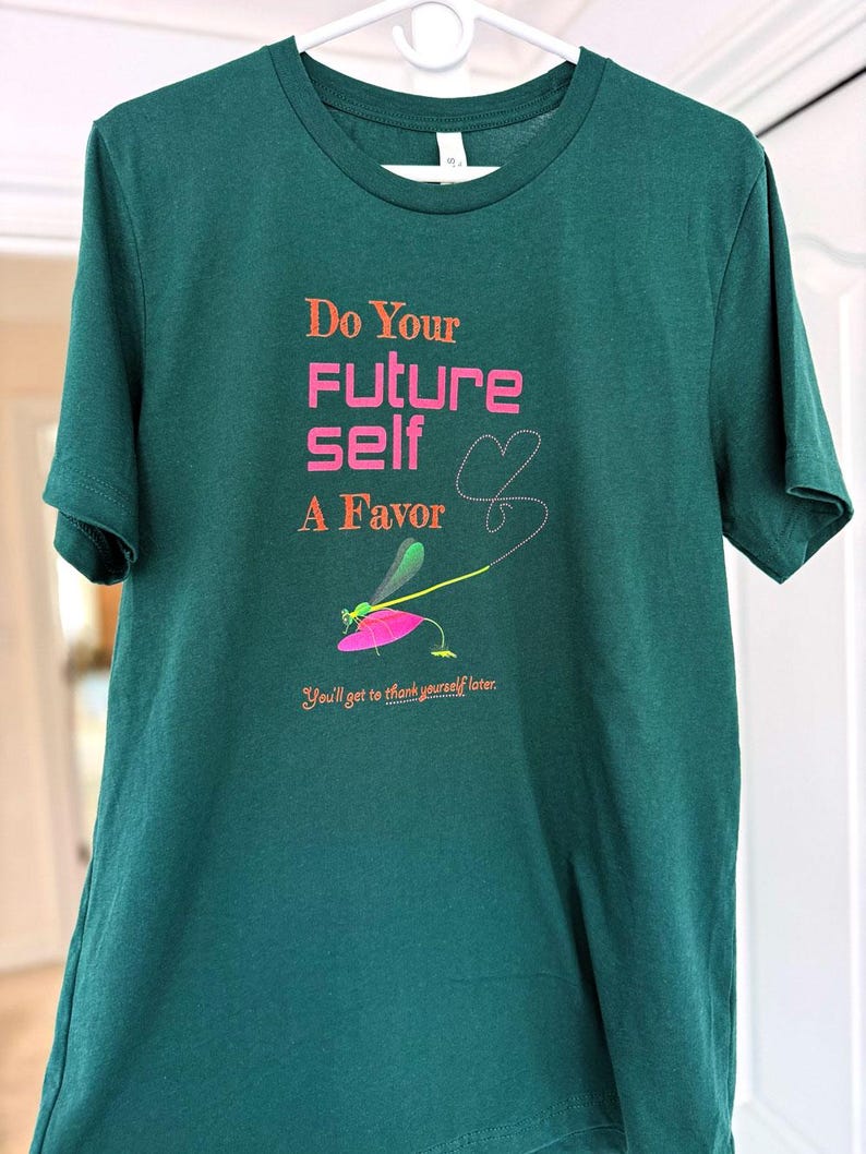 Puede incluir: Una camiseta verde oscuro con el texto "Do Your Future Self A Favor" en naranja y rosa. Un gr&aacute;fico de lib&eacute;lula est&aacute; debajo del texto, y la frase "You'll get to thank yourself later" est&aacute; impresa en la parte inferior.