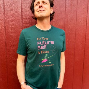 Puede incluir: Una camiseta verde oscuro con el texto "Do Your Future Self A Favor" en rosa y un gr&aacute;fico de lib&eacute;lula. La camiseta tambi&eacute;n incluye la frase "You'll get to thank yourself later". La persona lleva vaqueros azules.