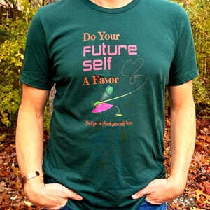 Puede incluir: Camiseta verde oscuro con el texto "Do Your Future Self A Favor" en rosa y naranja. Un gr&aacute;fico de lib&eacute;lula est&aacute; debajo del texto, con la frase "You'll get to thank yourself later." impresa debajo. La persona lleva vaqueros azules.