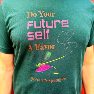 Puede incluir: Camiseta verde oscuro con el texto "Do Your Future Self A Favor" en naranja y rosa. Un gr&aacute;fico de lib&eacute;lula con un ala rosa y un cuerpo amarillo est&aacute; debajo del texto. La camiseta tambi&eacute;n tiene el texto "You'll get to thank yourself later."