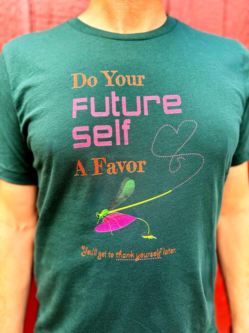 Puede incluir: Camiseta verde oscuro con el texto "Do Your Future Self A Favor" en naranja y rosa. Un gr&aacute;fico de lib&eacute;lula con un ala rosa y un cuerpo amarillo est&aacute; debajo del texto. La camiseta tambi&eacute;n tiene el texto "You'll get to thank yourself later."