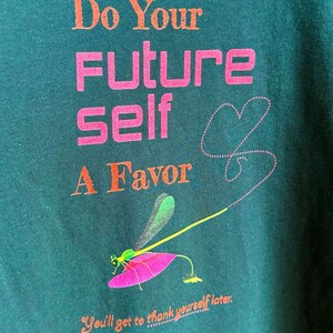 Puede incluir: Camiseta verde oscuro con el texto "Do Your Future Self A Favor" en naranja y rosa. Un gr&aacute;fico de lib&eacute;lula con alas verdes y una hoja rosa est&aacute; debajo del texto. La frase "You'll get to thank yourself later" est&aacute; en la parte inferior.