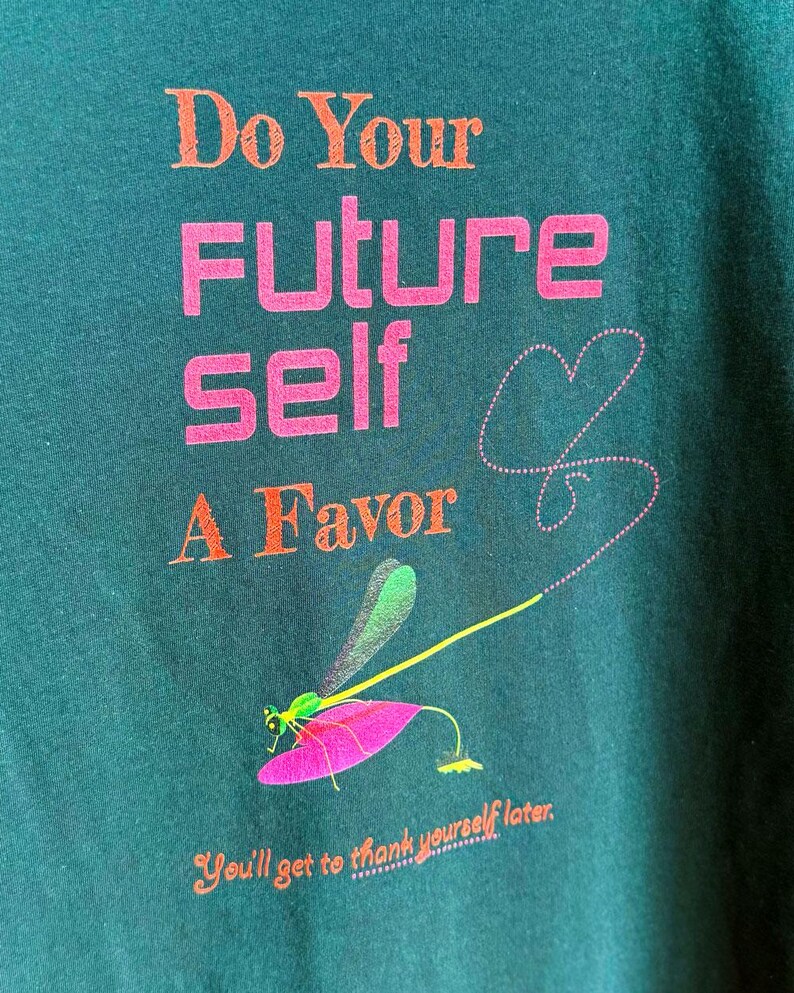 Puede incluir: Camiseta verde oscuro con el texto "Do Your Future Self A Favor" en naranja y rosa. Un gr&aacute;fico de lib&eacute;lula con alas verdes y una hoja rosa est&aacute; debajo del texto. La frase "You'll get to thank yourself later" est&aacute; en la parte inferior.