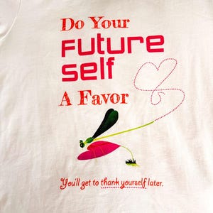 Puede incluir: Camiseta blanca con texto rojo: "Do Your FUTURE self A Favor". Ilustraci&oacute;n de lib&eacute;lula con detalles en rosa, verde y negro debajo del texto. La frase "You'll get to thank yourself later" est&aacute; impresa en rojo.