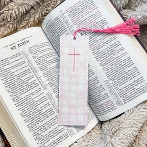 May include: A pink and white plaid bookmark with a pink cross and bunny silhouettes, accented with a pink tassel, rests inside an open Bible. The Bible pages display text.