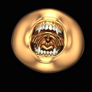 Puede incluir: Un objeto circular de tono dorado que presenta una boca muy abierta con dientes plateados, afilados y prominentes. La superficie metálica y lisa de los labios contrasta con el interior detallado y texturizado de la cavidad bucal, creando una forma audaz.
