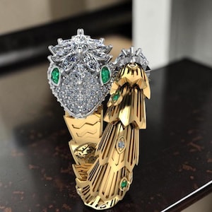 Könnte beinhalten: Ein gold-silberner Schlangenring mit detailliertem Design. Der Schlangenkopf ist silberfarben, besetzt mit kleinen, klaren Steinen und grünen Smaragdaugen. Der Körper ist goldfarben mit aufwendigen Mustern.