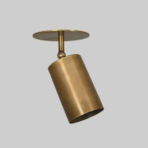 Könnte beinhalten: Ein goldfarbener, zylindrischer Deckenstrahler mit einer kreisförmigen Montageplatte. Der Strahler ist mit einem Kugelgelenk verstellbar und dient zur Lichtlenkung. Die Leuchte ist aus Metall und hat ein modernes Design.