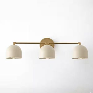 May include: A three-light vanity light fixture with a brass finish. The fixture has three off-white, dome-shaped shades. The light is mounted on a wall and is suitable for a bathroom or bedroom.