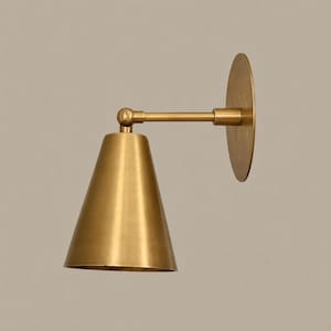 Puede incluir: Un aplique de pared con acabado en latón cepillado. La lámpara consta de una placa circular para la pared, un brazo horizontal y una pantalla cónica. Una bisagra visible conecta el brazo con la pantalla, permitiendo ajustar la posición.